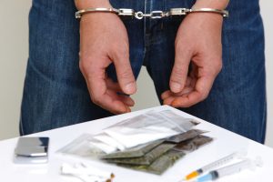 A person in handcuffs and blue jeans in front of a table filled with drugs and drug paraphernalia.