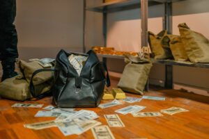 Money bags on the ground surrounded by hundred-dollar bills and gold bars.