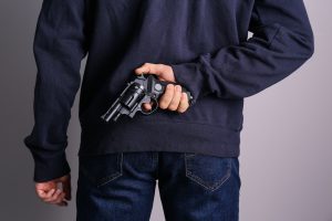 A man holding a gun behind his back wearing a dark blue sweatshirt and blue jeans