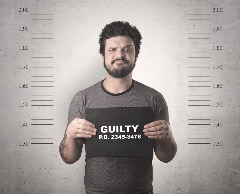 a man with a beard standing in a criminal lineup with a card that says, “Guilty.”