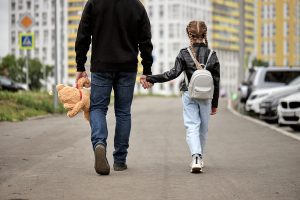 A man in a black jacket holds a teddy bear in one hand and the hand of a young girl in another.