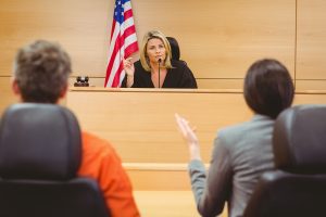 The backs of a man in an orange jump sitting next to a woman in a business suit. Both are facing a judge with a flag in the background.
