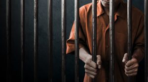 A person in an orange jumpsuit, from his lips to his waist, holds onto prison bars.